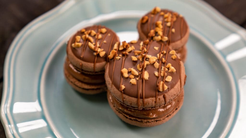A Mickey-shaped chocolate macaron topped with chopped nuts and chocolate drizzle sits on a plate during the Disney California Adventure Food & Wine Festival.