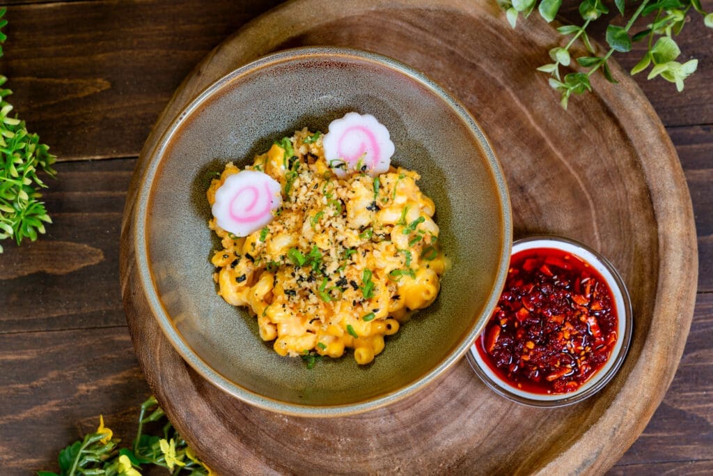 A bowl of ramen mac and cheese topped with crunchy crumbs and garnishes sits on a wooden serving tray with chili oil on the side.
