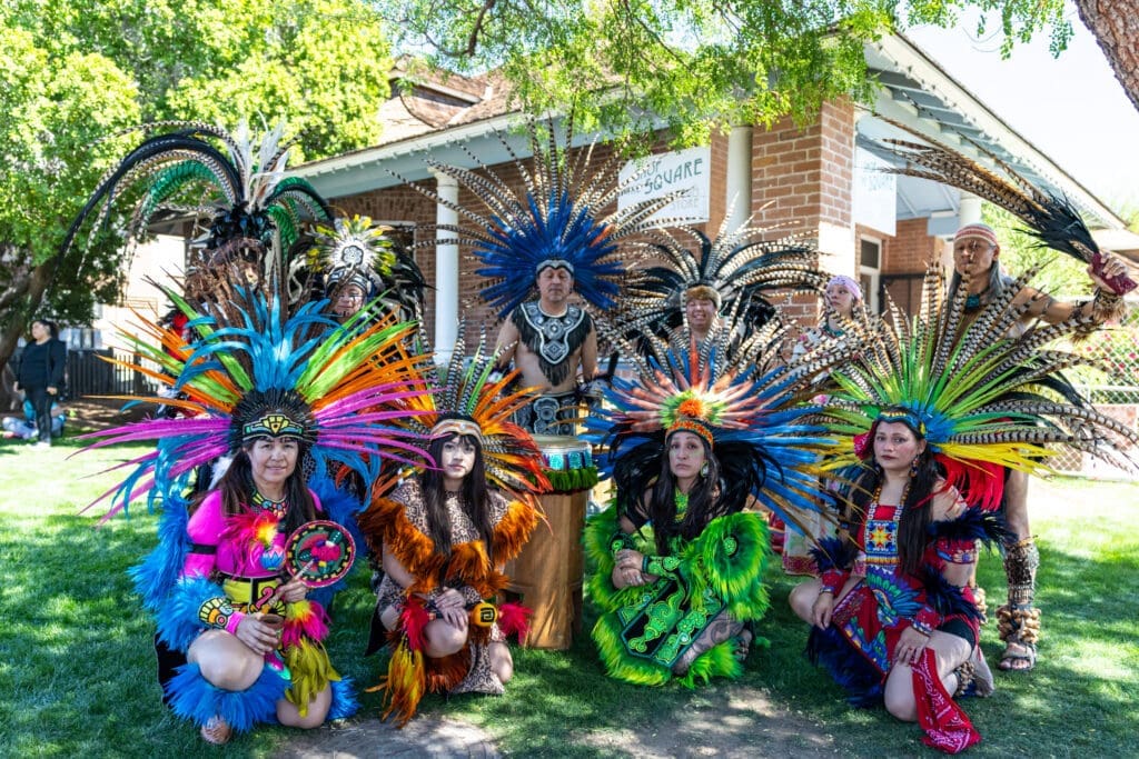 A group of cultural performers wearing bright feathered headdresses and traditional attire pose together on the lawn at Heritage Square during the Spirits of Mexico Festival.