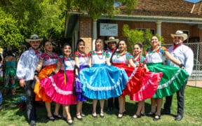 A group of folklórico dancers wearing brightly colored dresses and traditional attire pose together in front of a historic building at Heritage Square in Phoenix.