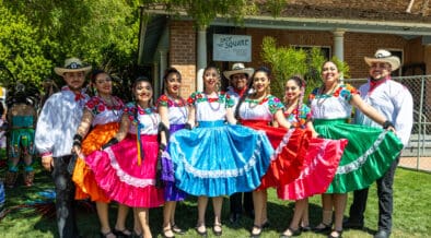 A group of folklórico dancers wearing brightly colored dresses and traditional attire pose together in front of a historic building at Heritage Square in Phoenix.