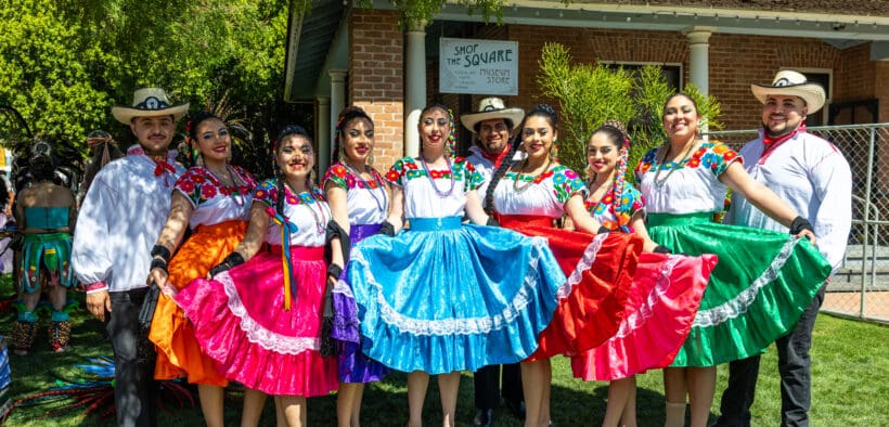 A group of folklórico dancers wearing brightly colored dresses and traditional attire pose together in front of a historic building at Heritage Square in Phoenix.