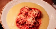 Meatballs topped with grated cheese and marinara sauce served over creamy polenta in a shallow bowl.
