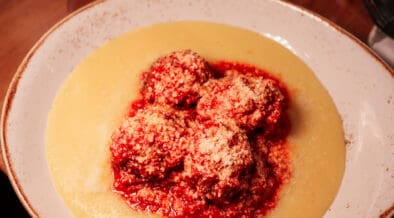 Meatballs topped with grated cheese and marinara sauce served over creamy polenta in a shallow bowl.