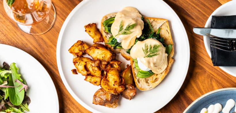 Overhead view of eggs benedict with hollandaise sauce, roasted potatoes and greens on a white plate at Apple Blossom's Easter brunch.