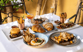 Table filled with brunch dishes including croissants, pancakes, burger, potatoes and cocktails in a shaded outdoor setting.