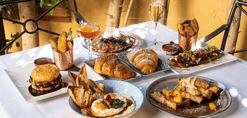 Table filled with brunch dishes including croissants, pancakes, burger, potatoes and cocktails in a shaded outdoor setting.