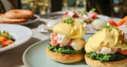 Lobster eggs Benedict topped with hollandaise sauce on an English muffin with sautéed greens, with oysters, avocado toast, and mimosas on a sunlit patio table.
