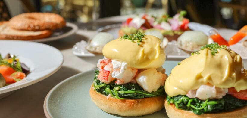 Lobster eggs Benedict topped with hollandaise sauce on an English muffin with sautéed greens, with oysters, avocado toast, and mimosas on a sunlit patio table.