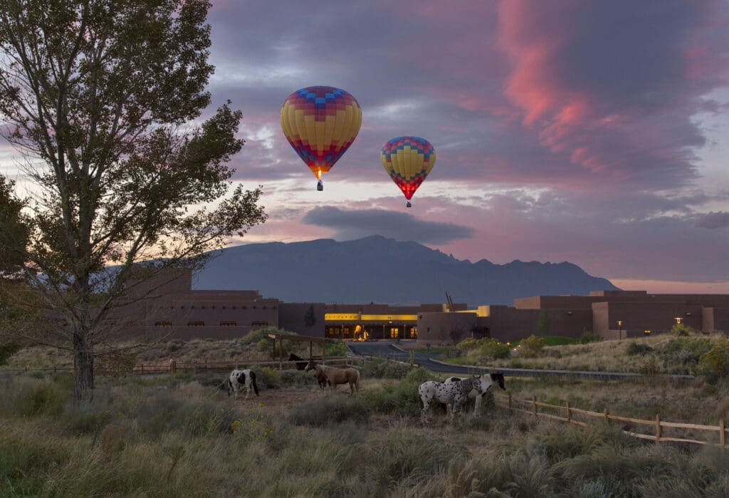 Two colorful hot air balloons rise above Hyatt Regency Tamaya Resort & Spa at sunrise with horses grazing in a fenced desert landscape and mountains in the background.