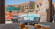 Poolside table with burgers, fries and drinks overlooking a resort pool with lounge chairs, umbrellas and adobe-style buildings in New Mexico.