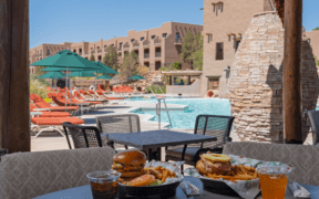 Poolside table with burgers, fries and drinks overlooking a resort pool with lounge chairs, umbrellas and adobe-style buildings in New Mexico.