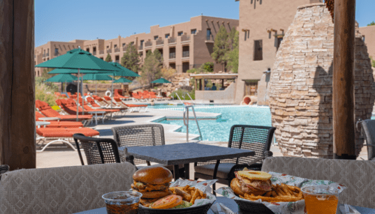 Poolside table with burgers, fries and drinks overlooking a resort pool with lounge chairs, umbrellas and adobe-style buildings in New Mexico.