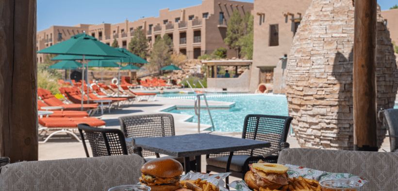Poolside table with burgers, fries and drinks overlooking a resort pool with lounge chairs, umbrellas and adobe-style buildings in New Mexico.