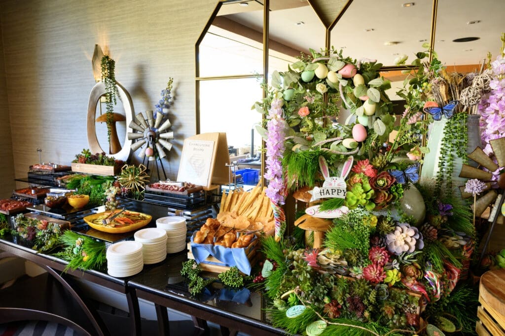 Easter brunch buffet with charcuterie, breads and decorative spring display featuring pastel eggs, flowers and greenery.