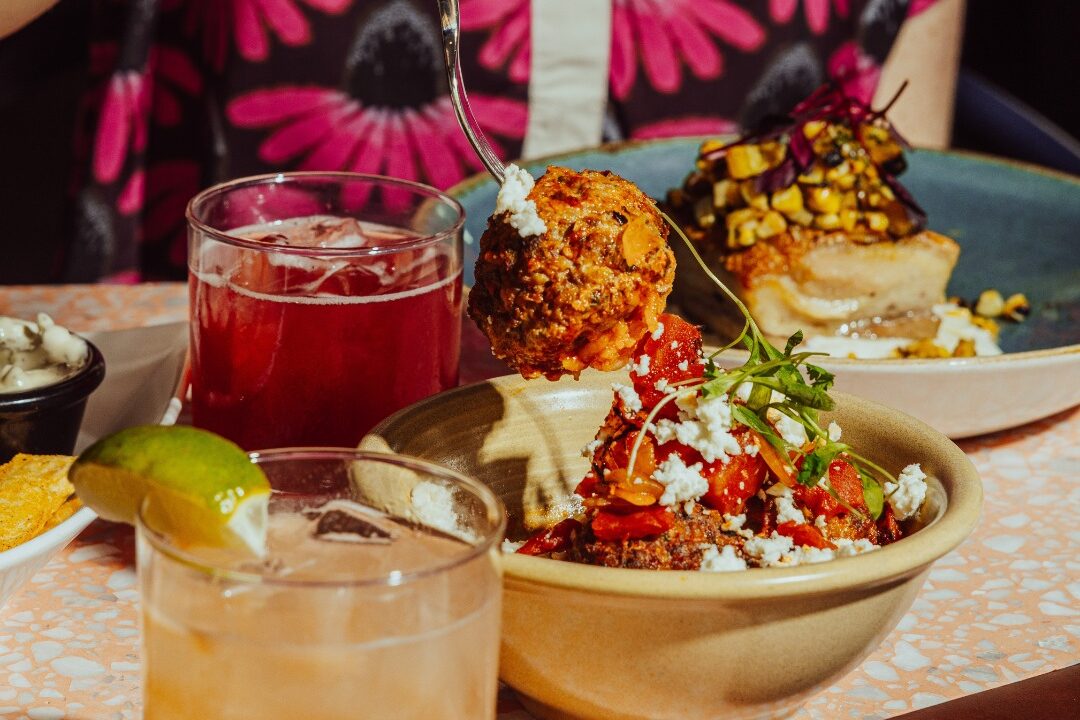 Person lifting a sauced meatball over a bowl topped with tomatoes and crumbled cheese, with cocktails and plated dishes at Kachina Cantina.