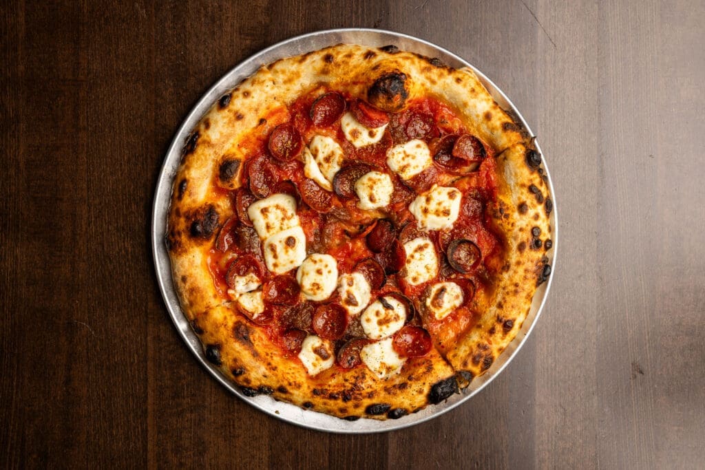 Overhead view of a wood-fired pepperoni pizza topped with melted mozzarella and tomato sauce on a metal tray from Little Johnny Bechamel's.