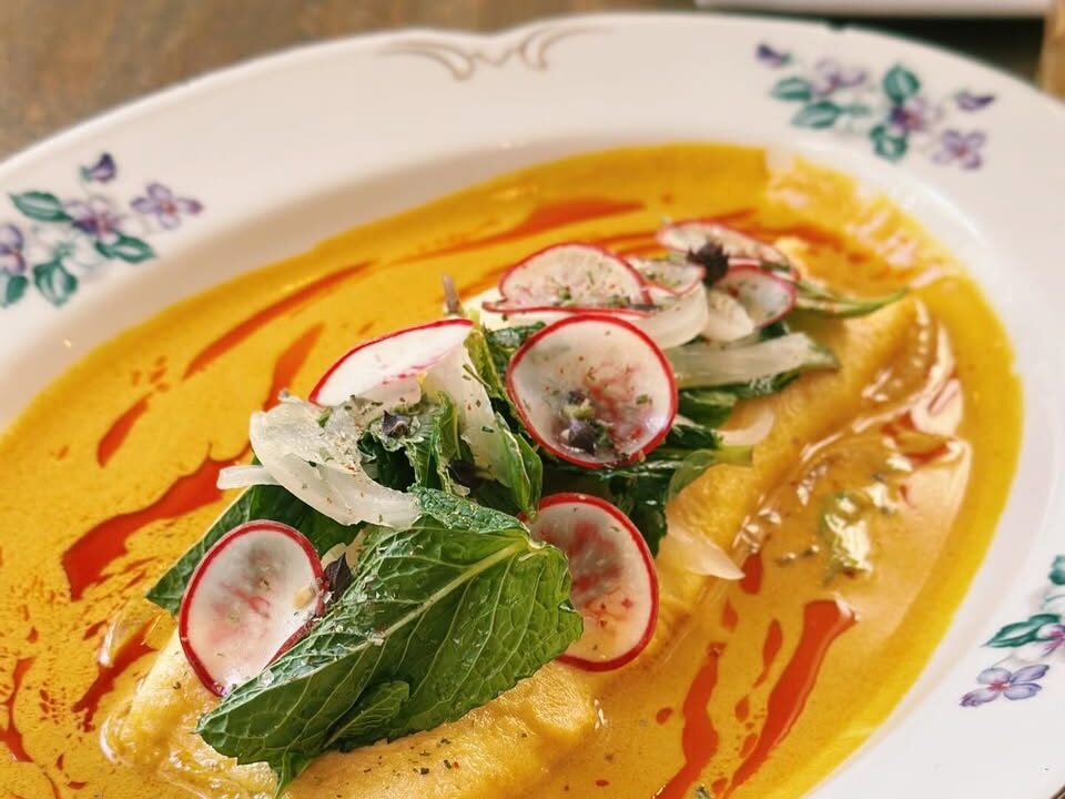 Elegant plated brunch dish featuring pasta or ravioli in a vibrant yellow sauce topped with fresh greens and thinly sliced radishes on a floral plate.