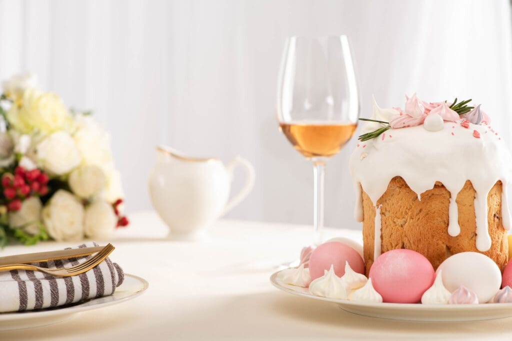 A selective focus of delicious Easter cake decorated with meringue with pink and white eggs on plate near wine glass and flowers.