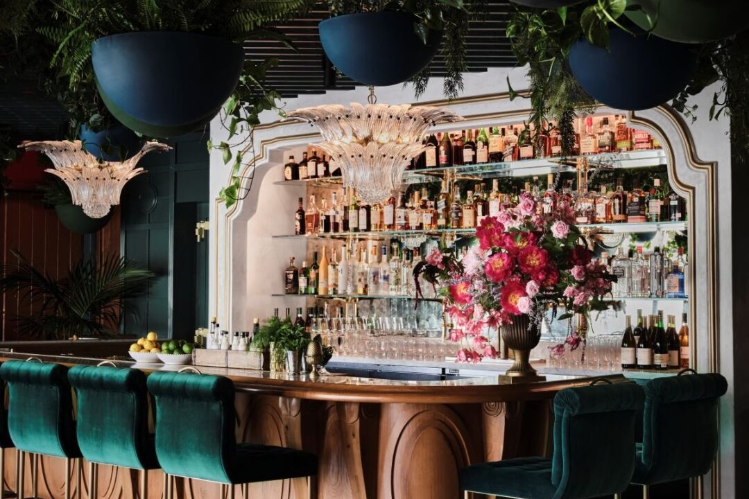 Elegant bar interior with green velvet stools, floral arrangement and hanging plants creating a lush garden-inspired atmosphere.