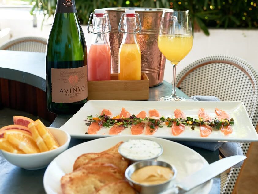 Brunch table with smoked salmon, toast, citrus slices and mimosas served with a bottle of sparkling wine and fresh juices in a bright patio setting.