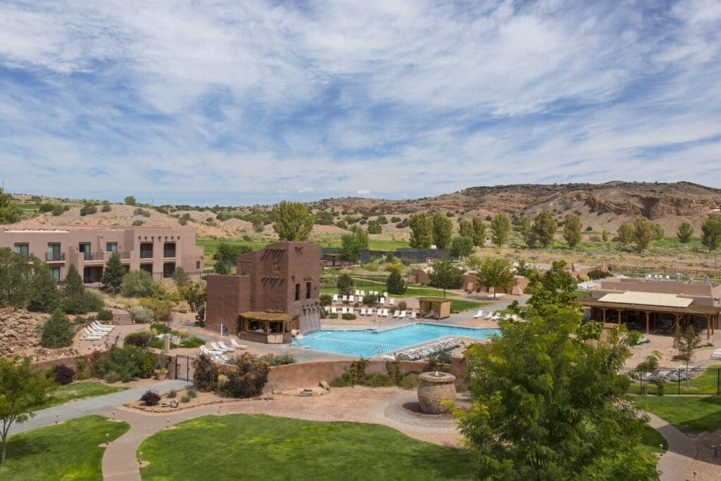 Outdoor resort pool with lounge chairs, adobe-style buildings and landscaped grounds set against a desert backdrop in New Mexico.