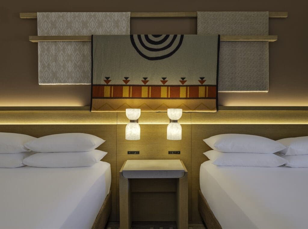 Two queen beds with white linens in a guestroom at Hyatt Regency Tamaya Resort & Spa featuring a Native-inspired woven wall hanging, wood headboard and soft ambient lighting.