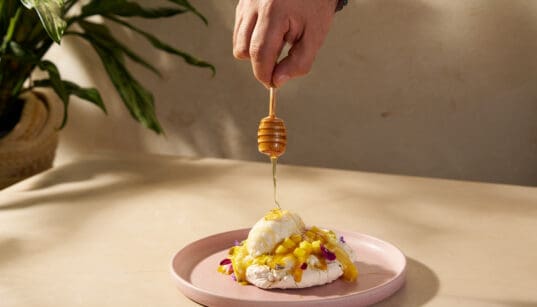 Honey being drizzled over the Abejas Pavlova dessert with mango, whipped cream and coconut ice cream.