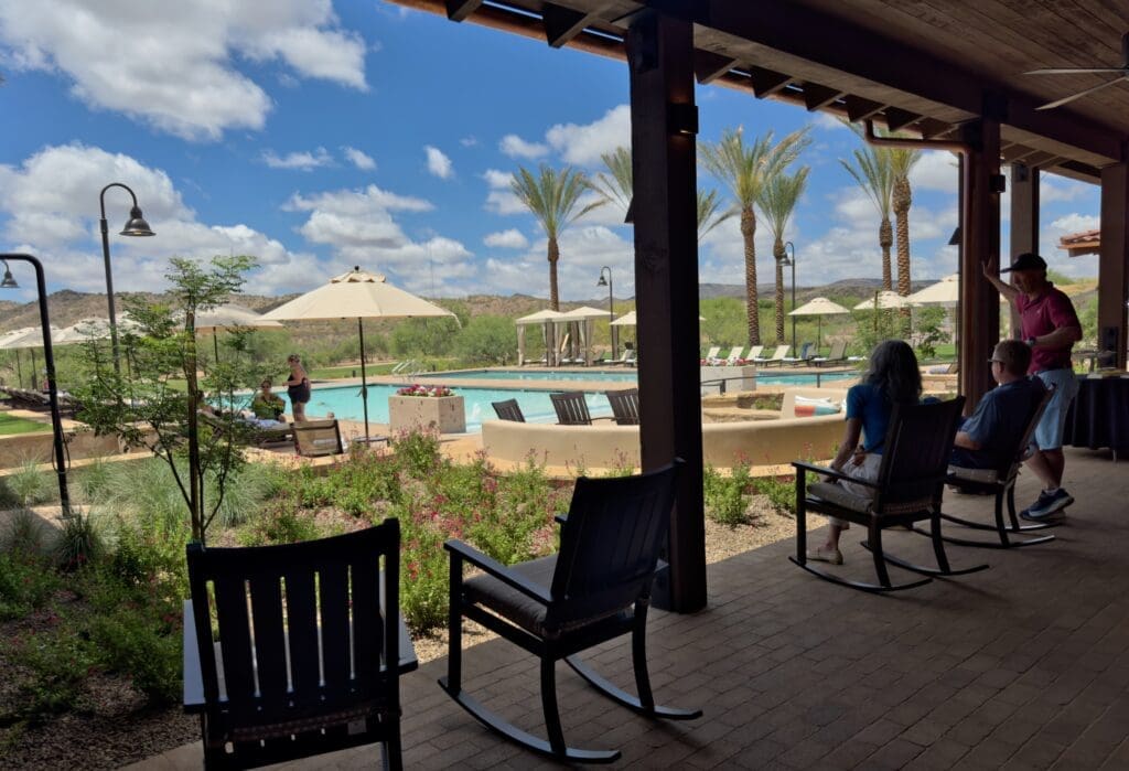 Guests sitting in rocking chairs on a shaded patio overlooking the pool area at Rancho de los Caballeros resort in Wickenburg, Arizona.