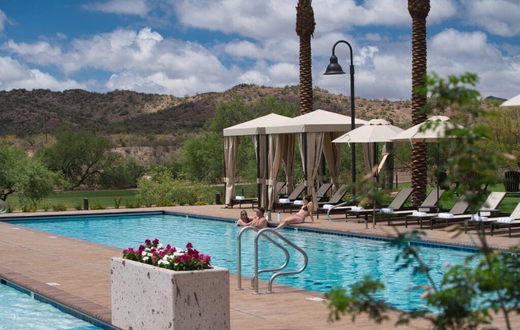 Guests relaxing by the pool and cabanas at Rancho de los Caballeros resort with desert hills and palm trees in the background.