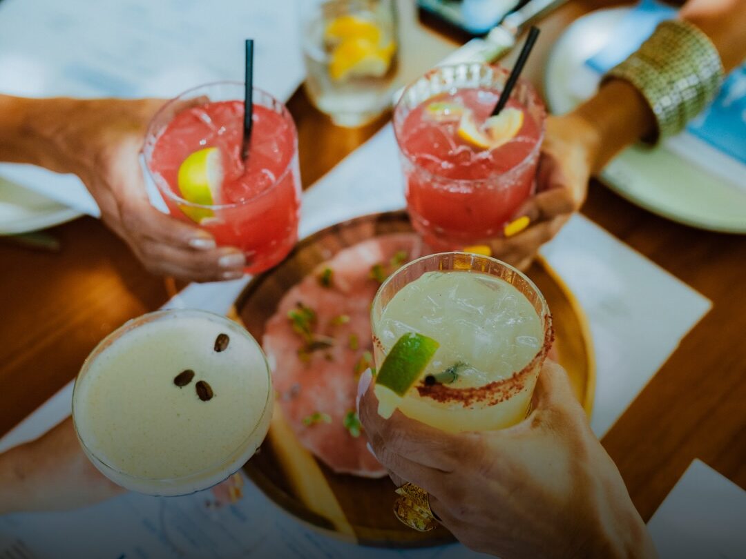 Four people clinking cocktails over a table with small plates of food, including a citrus margarita, pink cocktails, and a creamy espresso martini, during a lively brunch setting.