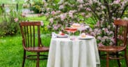 Intimate outdoor Easter table for two set beneath blooming pink trees with desserts and drinks on a white tablecloth.