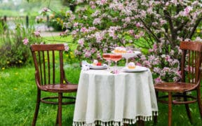 Intimate outdoor Easter table for two set beneath blooming pink trees with desserts and drinks on a white tablecloth.