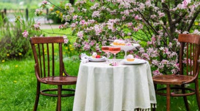 Intimate outdoor Easter table for two set beneath blooming pink trees with desserts and drinks on a white tablecloth.