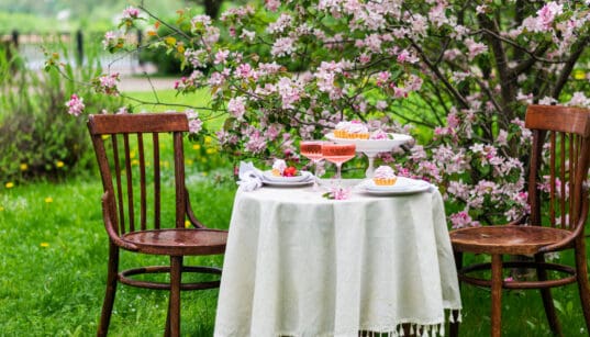 Intimate outdoor Easter table for two set beneath blooming pink trees with desserts and drinks on a white tablecloth.