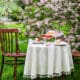 Intimate outdoor Easter table for two set beneath blooming pink trees with desserts and drinks on a white tablecloth.
