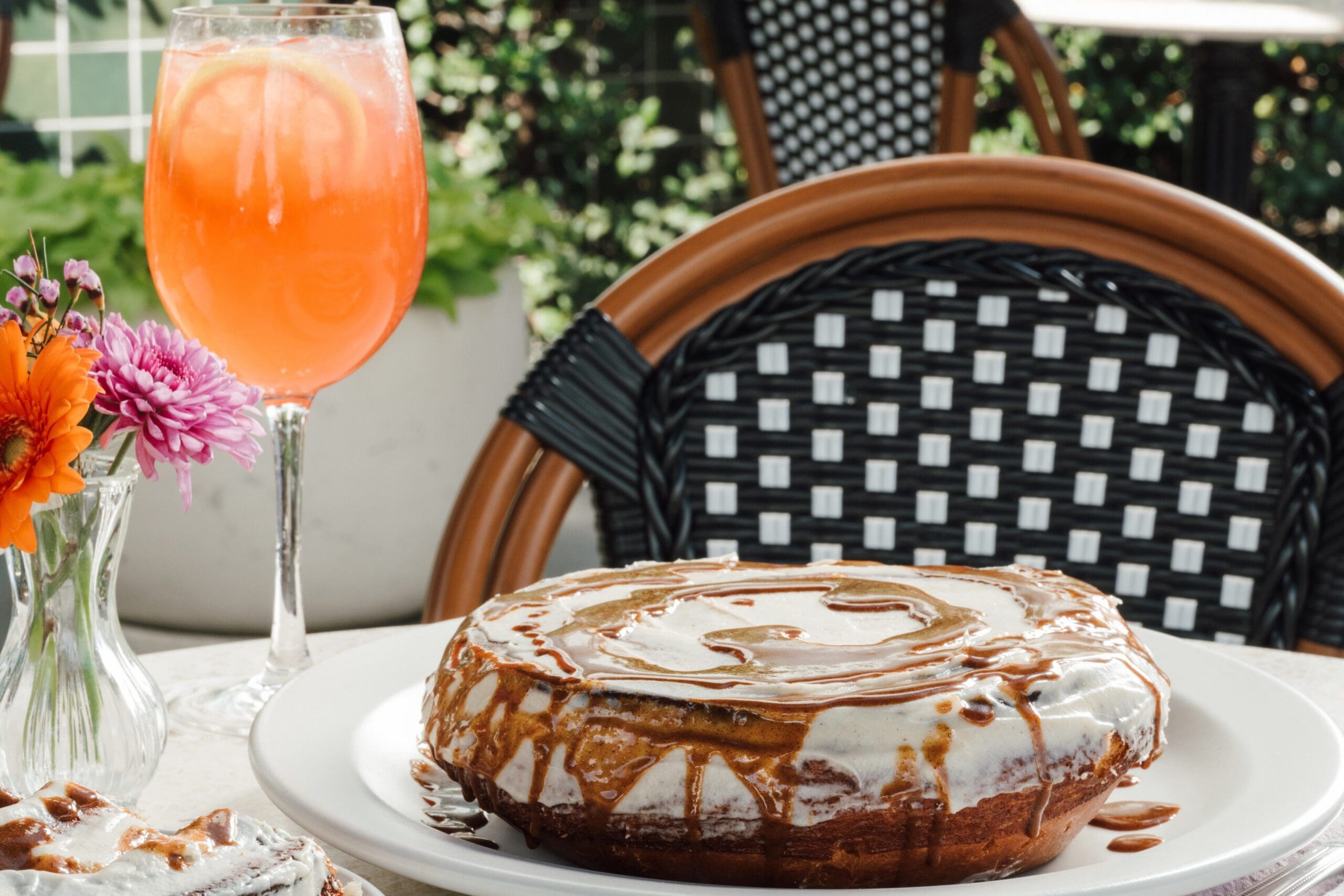 Iced cinnamon roll on a plate with a cocktail and flowers on an outdoor café table at The Henry.