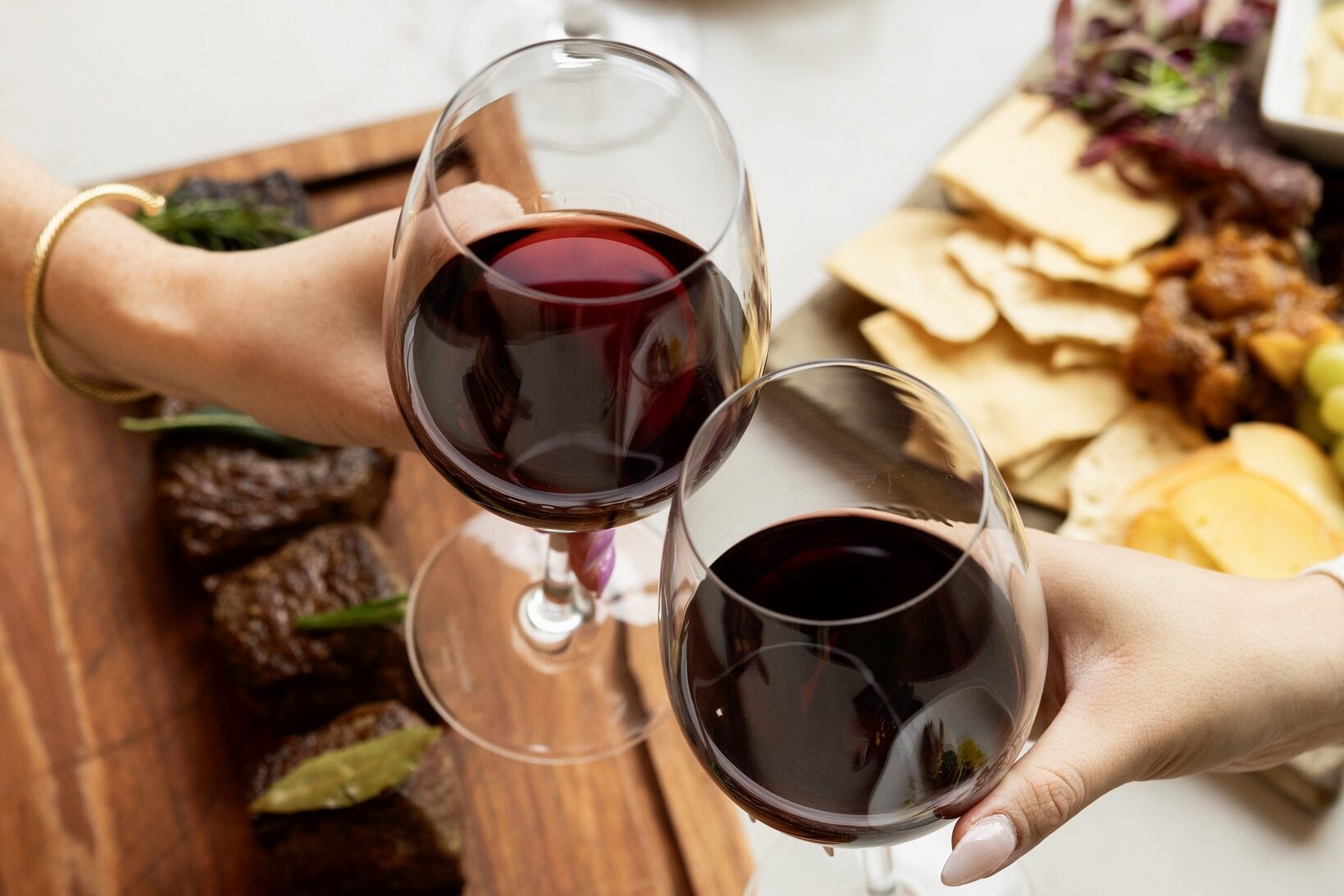 Two hands clinking glasses of red wine over a wooden board with steak bites, alongside a charcuterie spread and plated dishes at Urban Farmer in Denver.
