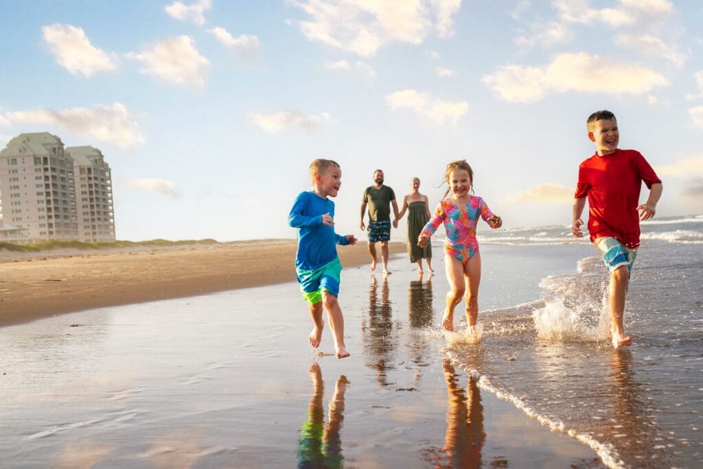 Children running and playing in the ocean waves with parents walking behind them on South Padre Island beach.