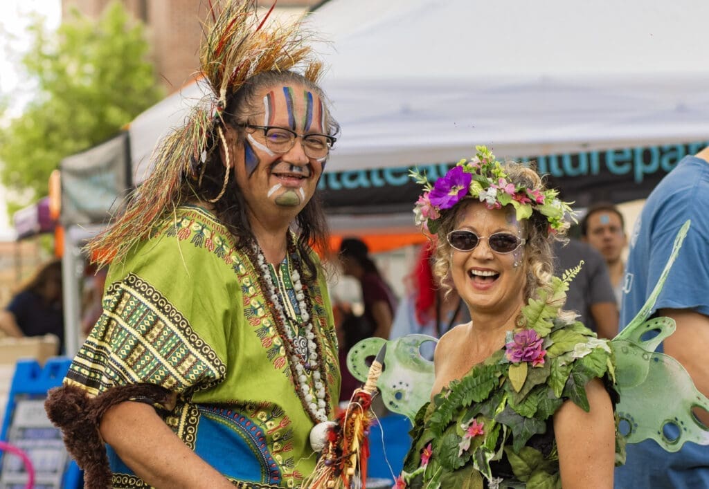Two adults in colorful costumes and face paint smiling at a Flagstaff festival booth, one wearing a green outfit and the other dressed like a fairy.
