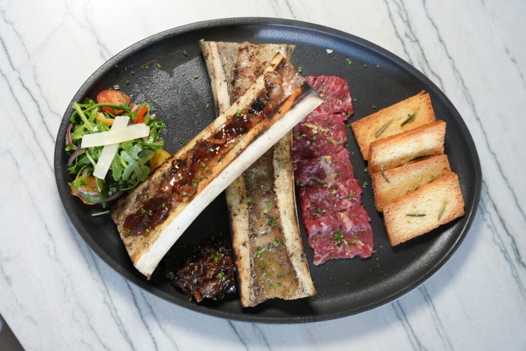 Bone marrow on a plate with mixed arugula salad, sliced steak and rectangular croutons at Jack’s Modern Steakhouse Lone Tree.