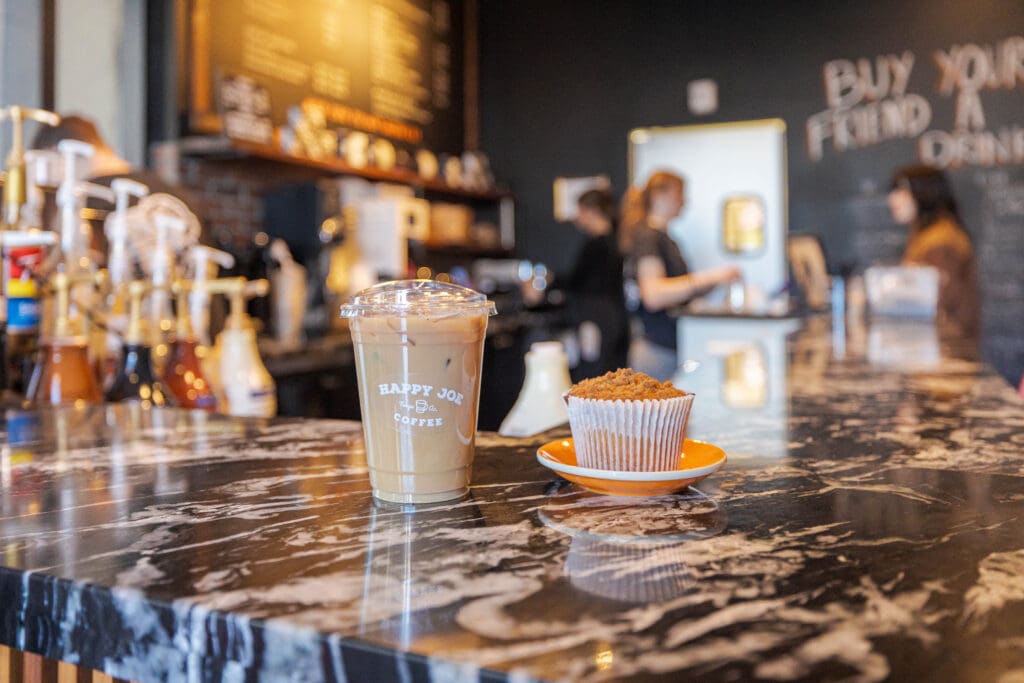 Iced coffee and a muffin on a marble counter inside a coffee shop with baristas in the background.