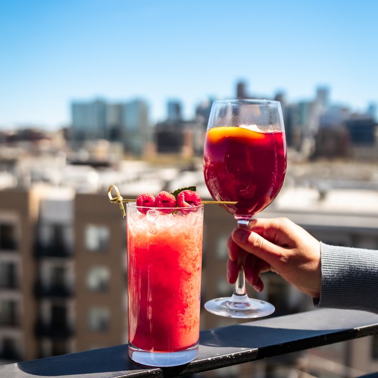 Two vibrant brunch cocktails, including a spritz and sparkling wine, held against a blurred Denver skyline from a rooftop restaurant.