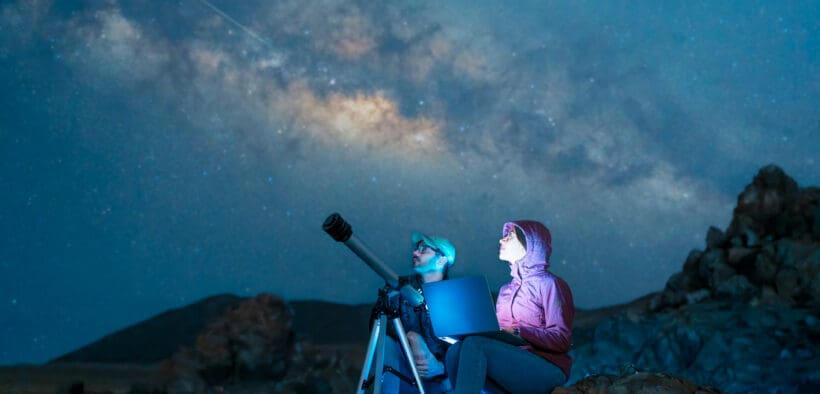 Couple sitting in the desert watching the stars and Milky Way next to a telescope in Utah.