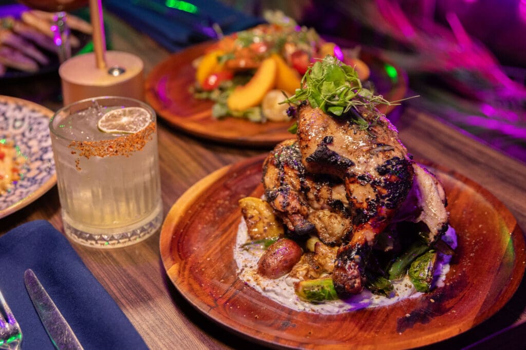 A grilled chicken entree with roasted vegetables and cocktail at upscale El Paso restaurant with colorful lighting.