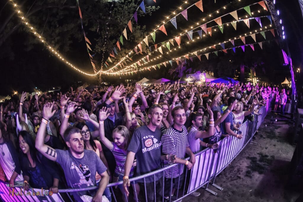 Large crowd gathered at an outdoor concert at night with string lights and colorful flags overhead at Flagstaff Hullabaloo Festival.
