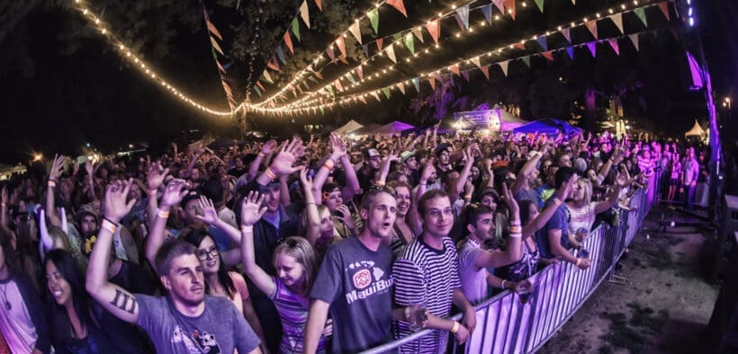 Large crowd gathered at an outdoor concert at night with string lights and colorful flags overhead at Flagstaff Hullabaloo Festival.