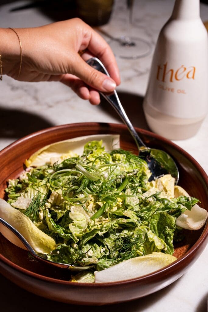 Green salad with herbs and Théa olive oil bottle on table at Mediterranean rooftop restaurant.