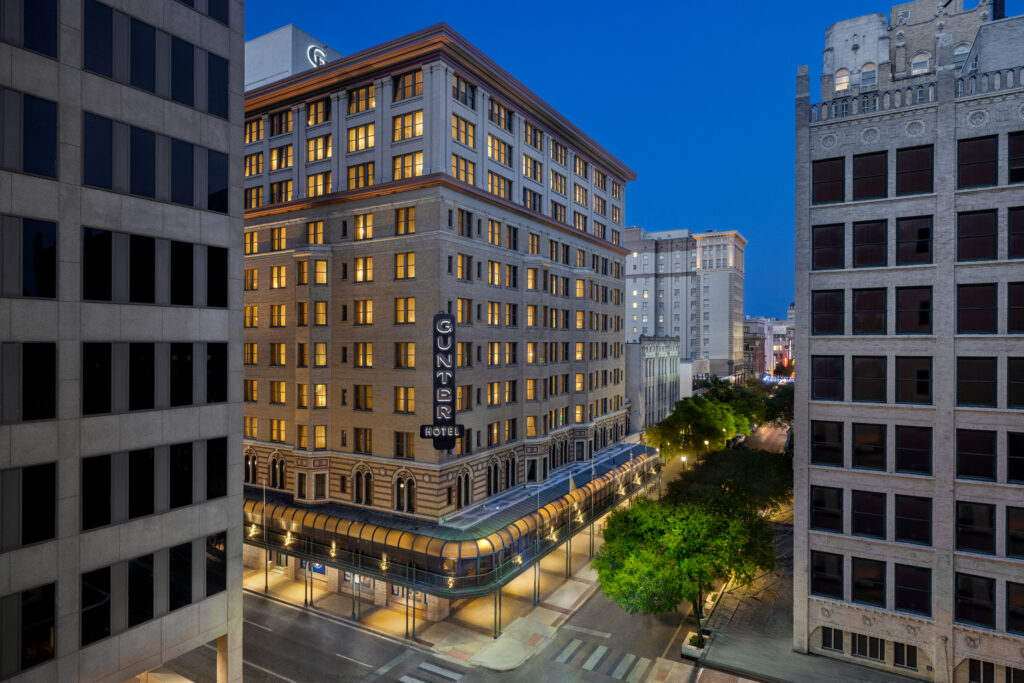Exterior of Gunter Hotel in downtown San Antonio at dusk with illuminated windows and city buildings surrounding.