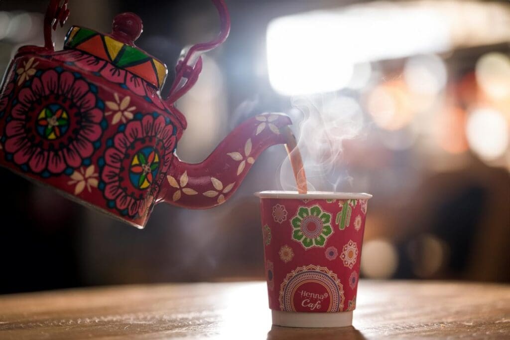 Decorative teapot pouring hot chai into a patterned paper cup with steam rising in a café setting.